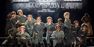 A group of boys in ragged clothing stand together on stage in front of a building labeled "God Is Love Home for Paupers and Orphans," appearing to sing or shout.